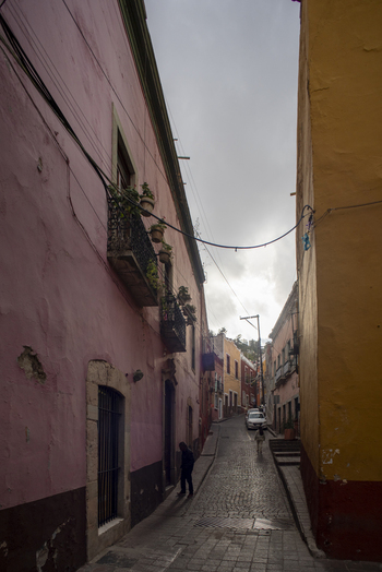 View of a narrow cobblestone street in Guanajuato featuring colorful colonial architecture with pink and yellow walls balconies with potted plants and a person walking in Guanajuato Guanajuato Mexico. View of a narrow cobblestone street in Guanajuato featuring colorful colonial architecture with pink and yellow walls balconies with potted plants and a person walking in Guanajuato Guanajuato Mexico.