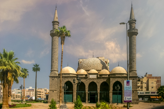 View of the historic Al Anbariya Masjid featuring stone masonry multiple domes and tall minarets framed by palm trees in Madinah Al Madinah Province Saudi Arabia.