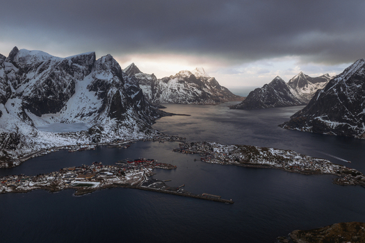 Aerial view of snowy mountain peaks rise majestically from the dark still waters surrounding the quaint village of Reinebringen Nordland Norway.