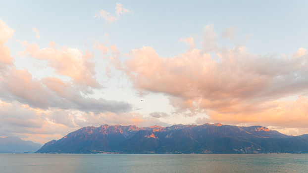View of the Alps reflecting in the calm waters of Lake Geneva under a soft sunset sky with pink and orange clouds in Bourg en Lavaux Vaud Switzerland. View of the Alps reflecting in the calm waters of Lake Geneva under a soft sunset sky with pink and orange clouds in Bourg en Lavaux Vaud Switzerland.