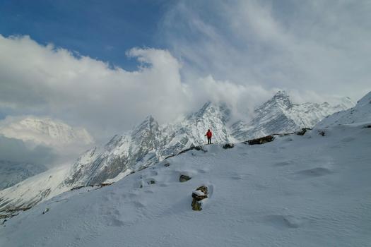 Aerial view of a hiker in a red jacket standing on a snow covered mountain ridge with the majestic Himalayan peaks and clouds in the background in Nepal.