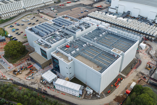 Aerial view of a large modern data center facility with industrial cooling units on the roof surrounding parking area and construction equipment in the United Kingdom.