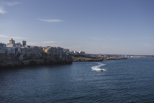View of the white buildings of the historic town perched on rocky cliffs overlooking the blue Adriatic Sea with a boat wake Polignano a Mare Apulia Italy.