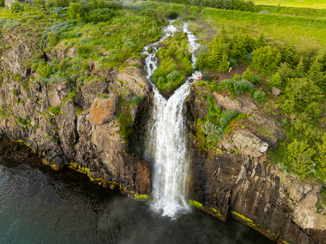Aerial view of a waterfall cascading down a rocky cliff into the sea surrounded by lush green vegetation in Akureyri Akureyrarbaer Iceland.