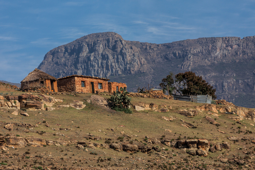 View of traditional stone and mud houses with thatched roofs situated on a rocky hillside against a backdrop of rugged mountains in South Africa.