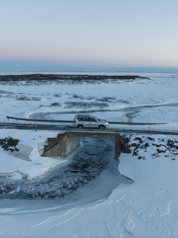 Aerial view of a silver SUV driving across a small stone bridge over a partially frozen river in a vast snow covered landscape in Kirkjubaejarklaustur Skaftarhreppur Iceland. Aerial view of a silver SUV driving across a small stone bridge over a partially frozen river in a vast snow covered landscape in Kirkjubaejarklaustur Skaftarhreppur Iceland.