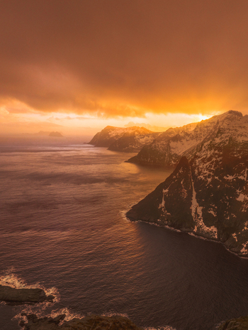 Aerial view of jagged cliffs plunge into the dark waters under a fiery clouded sky Nordland Norway.