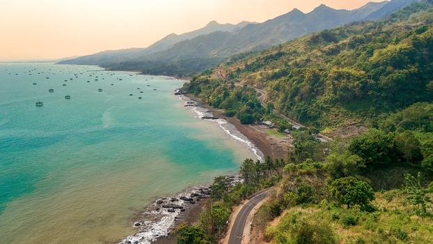 Aerial view of a winding coastal road along a dark sand beach with turquoise sea fishing platforms and lush green hills in Sukabumi Indonesia. Aerial view of a winding coastal road along a dark sand beach with turquoise sea fishing platforms and lush green hills in Sukabumi Indonesia.