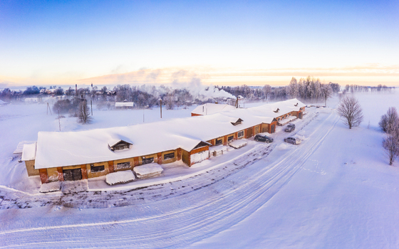 Aerial view of a long building covered in snow in a rural landscape with trees and smoke rising in the distance Latvia. Aerial view of a long building covered in snow in a rural landscape with trees and smoke rising in the distance Latvia.