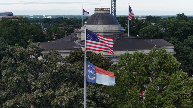 Aerial view of the North Carolina State Capitol with the American flag and North Carolina state flag waving in the foreground surrounded by trees in Raleigh North Carolina United States. Aerial view of the North Carolina State Capitol with the American flag and North Carolina state flag waving in the foreground surrounded by trees in Raleigh North Carolina United States.
