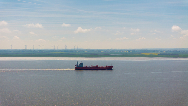 Brunsbuttel Germany 30 May 2025 Aerial view of a red cargo ship navigating the Elbe river with wind turbines visible on the distant green coastline. Brunsbuttel Germany 30 May 2025 Aerial view of a red cargo ship navigating the Elbe river with wind turbines visible on the distant green coastline.