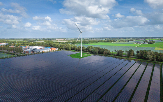 Aerial view of a large solar farm featuring a prominent wind turbine situated in a rural landscape with green fields and industrial buildings in Warmenhuizen North Holland Netherlands.