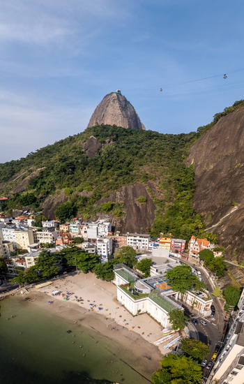 Aerial view of Sugarloaf Mountain rising above Praia da Urca beach with people on the sand and urban buildings nestled against green hills Rio de Janeiro State of Rio de Janeiro Brazil.