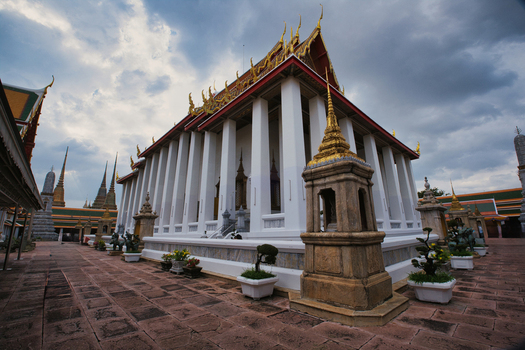 View of The Grand Palace featuring ornate gold roof architecture and white columns set against a cloudy sky in Bangkok Thailand. View of The Grand Palace featuring ornate gold roof architecture and white columns set against a cloudy sky in Bangkok Thailand.