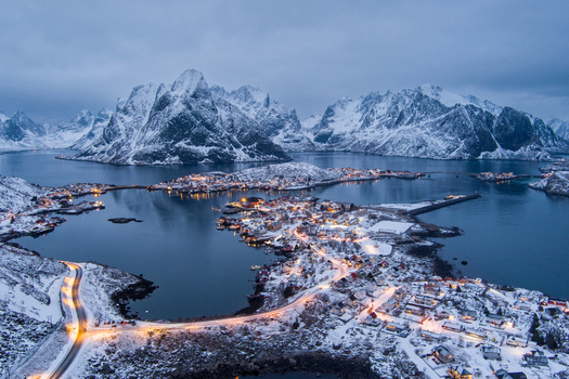 Aerial view of the snowy coastal village of Lofoten with illuminated roads and houses nestled between dramatic mountains and the sea in Nordland Norway.
