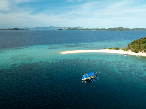 Aerial view of Malcapuya Island with a traditional outrigger boat in clear turquoise waters surrounded by coral reefs and distant islands in Coron Mimaropa Philippines. Aerial view of Malcapuya Island with a traditional outrigger boat in clear turquoise waters surrounded by coral reefs and distant islands in Coron Mimaropa Philippines.