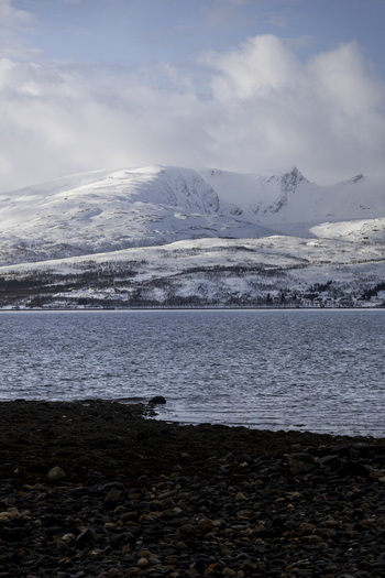 View of snow capped mountains rising above the calm waters of a fjord with a rocky shoreline under a cloudy sky in Kvaloya Troms Norway. View of snow capped mountains rising above the calm waters of a fjord with a rocky shoreline under a cloudy sky in Kvaloya Troms Norway.