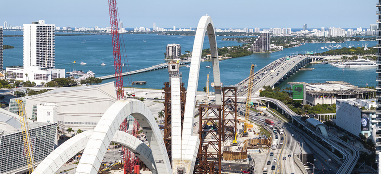 Aerial view of cranes towering over the construction of a grand archway for Signature Bridge near the Kaseya Center contrasted against the bright blue waters of Biscayne Bay Miami Florida USA. Aerial view of cranes towering over the construction of a grand archway for Signature Bridge near the Kaseya Center contrasted against the bright blue waters of Biscayne Bay Miami Florida USA.