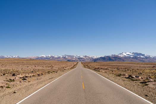View of the Mirador de los Andes road stretching through the arid high altitude landscape towards snow capped mountains under a clear blue sky in Caylloma Arequipa Peru.