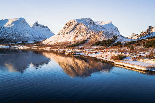 Aerial view of snowy mountains meet the serene reflective waters under a soft pastel sky Indregård Troms Norway. Aerial view of snowy mountains meet the serene reflective waters under a soft pastel sky Indregård Troms Norway.