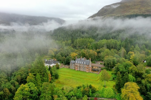 Aerial view of a historic stone manor house nestled in a lush green valley surrounded by dense forests and misty mountains in Glencoe Scotland United Kingdom.