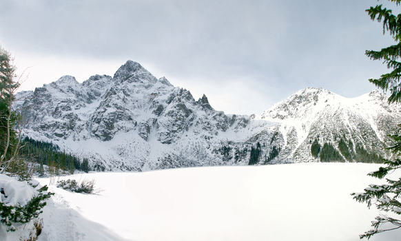 View of the snow covered peaks of the Tatra Mountains overlooking the frozen Morskie Oko lake surrounded by pine trees in Tatra County Lesser Poland Voivode Poland. View of the snow covered peaks of the Tatra Mountains overlooking the frozen Morskie Oko lake surrounded by pine trees in Tatra County Lesser Poland Voivode Poland.