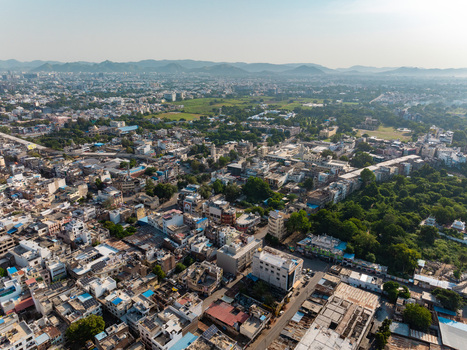 Aerial view of the dense urban sprawl featuring residential rooftops commercial buildings scattered green trees and distant hills under a clear sky in Udaipur Rajasthan India.