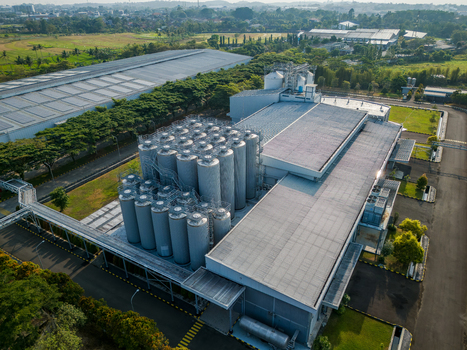 Aerial view of an industrial facility featuring a cluster of large metal storage silos and a modern factory building surrounded by green fields in Semarang Regency Central Java Indonesia. Aerial view of an industrial facility featuring a cluster of large metal storage silos and a modern factory building surrounded by green fields in Semarang Regency Central Java Indonesia.