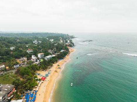 Aerial view of the tropical coastline with a sandy beach turquoise ocean water and lush green palm trees surrounding coastal buildings Dodanduwa Southern Province Sri Lanka. Aerial view of the tropical coastline with a sandy beach turquoise ocean water and lush green palm trees surrounding coastal buildings Dodanduwa Southern Province Sri Lanka.