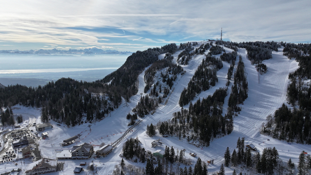 Aerial view of snow covered slopes descend from a peak crowned with a telecommunications tower contrasting with the dark evergreens and distant mountains Gex Auvergne Rhône Alpes France. Aerial view of snow covered slopes descend from a peak crowned with a telecommunications tower contrasting with the dark evergreens and distant mountains Gex Auvergne Rhône Alpes France.