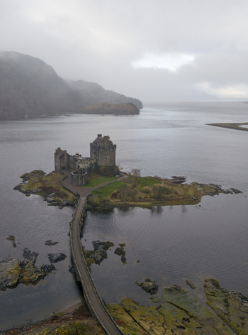 Aerial view of Eilean Donan Castle and its iconic stone bridge over the calm waters of Loch Duich with misty mountains in the background Dornie Scotland United Kingdom.