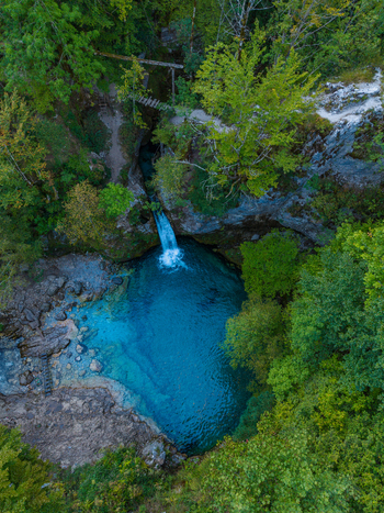 Aerial view of the Blue Eye Theth a vibrant turquoise natural pool fed by a waterfall surrounded by dense green forest and rocky terrain in Theth Shkoder County Albania. Aerial view of the Blue Eye Theth a vibrant turquoise natural pool fed by a waterfall surrounded by dense green forest and rocky terrain in Theth Shkoder County Albania.