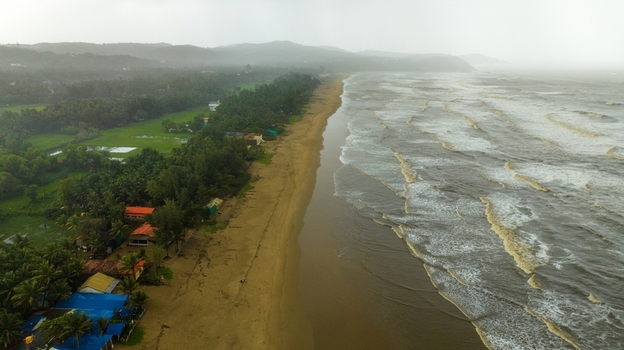 Aerial view of Gokarna beach with dark stormy waves crashing on the sandy shore lush green palm trees and coastal buildings under a cloudy overcast sky in Gokarna Karnataka India.