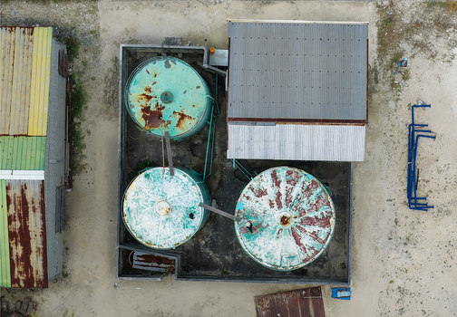 Aerial view of three weathered storage tanks with rusted tops and corrugated metal roofs on sandy ground Hanimaadhoo Haa Dhaalu Atoll Maldives.