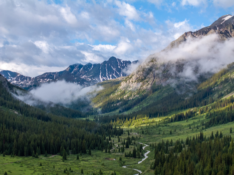 View of a lush mountain valley with a winding stream dense evergreen forests and misty snow capped peaks under a cloudy sky in Colorado United States.