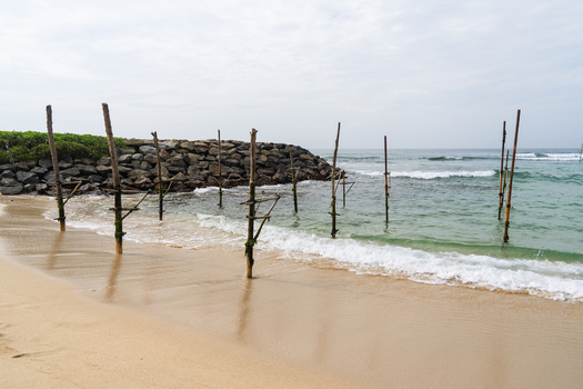 View of traditional stilt fishing poles in the shallow turquoise waters along a sandy beach with a stone breakwater under a cloudy sky in Koggala Southern Province Sri Lanka. View of traditional stilt fishing poles in the shallow turquoise waters along a sandy beach with a stone breakwater under a cloudy sky in Koggala Southern Province Sri Lanka.