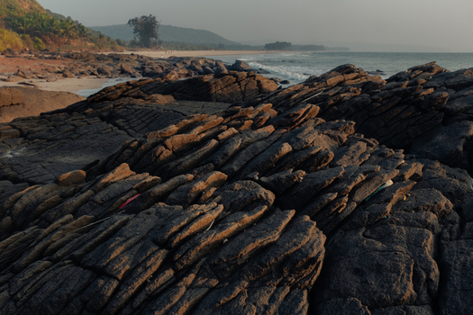 View of rugged dark rocks meet the shoreline as gentle waves roll onto a sandy beach framed by distant hills under a soft sky Chiplun Maharashtra India. View of rugged dark rocks meet the shoreline as gentle waves roll onto a sandy beach framed by distant hills under a soft sky Chiplun Maharashtra India.