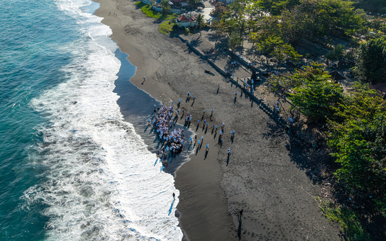 Aerial view of the Ngelarung Sea Offering Ceremony at Pura Segara Goa Lawah with people in traditional white clothing gathered on the black sand beach in Dawan Bali Indonesia.
