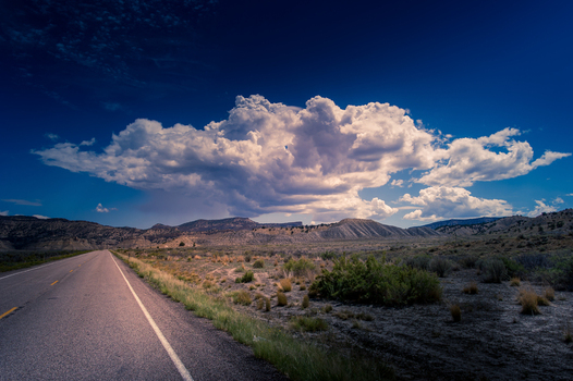 View of a road stretching through the desert landscape of Bryce Canyon National Park with distant mountains and a large cloud formation under a deep blue sky in Utah United States.