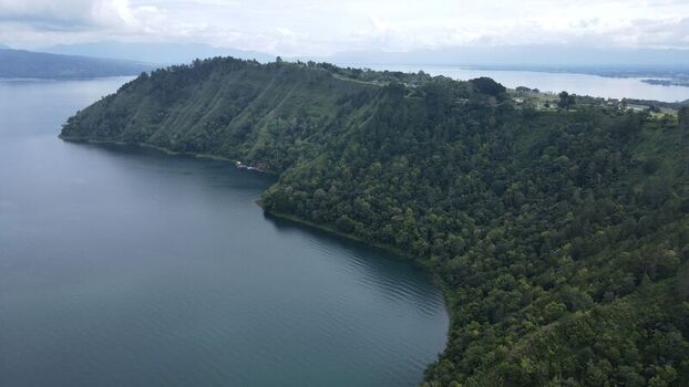 Aerial view of Tara Bunga hill covered in lush vegetation extending into the deep blue waters of Lake Toba under a cloudy sky in Tampahan North Sumatra Indonesia. Aerial view of Tara Bunga hill covered in lush vegetation extending into the deep blue waters of Lake Toba under a cloudy sky in Tampahan North Sumatra Indonesia.