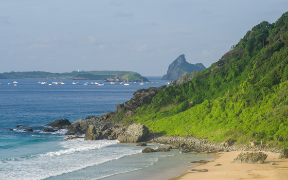 View of the sandy beach and turquoise ocean with lush green hills and rocky cliffs at Ilha de Fernando de Noronha in Fernando de Noronha State of Pernambuco Brazil. View of the sandy beach and turquoise ocean with lush green hills and rocky cliffs at Ilha de Fernando de Noronha in Fernando de Noronha State of Pernambuco Brazil.