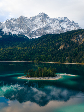 Aerial view of the turquoise Eibsee lake featuring a small forested island with the snow capped Zugspitze mountain and dense alpine forests in Grainau Bavaria Germany. Aerial view of the turquoise Eibsee lake featuring a small forested island with the snow capped Zugspitze mountain and dense alpine forests in Grainau Bavaria Germany.