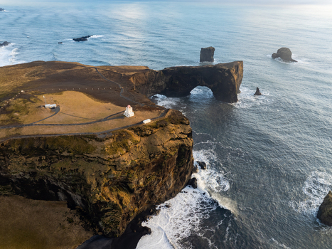 Aerial view of Dyrholaey arch with its iconic lighthouse rugged cliffs and crashing waves along the coastline in Myrdalshreppur Iceland. Aerial view of Dyrholaey arch with its iconic lighthouse rugged cliffs and crashing waves along the coastline in Myrdalshreppur Iceland.