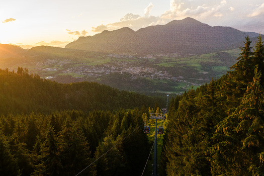 Aerial view of Fiemme Valley at sunset with a cable car gliding over dense pine forests and mountain slopes in Trentino Alto Adige Italy.