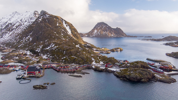 Aerial view of Lofoten featuring traditional red cabins snowy mountains and fish farming cages along the rocky coastline in Nordland Norway.