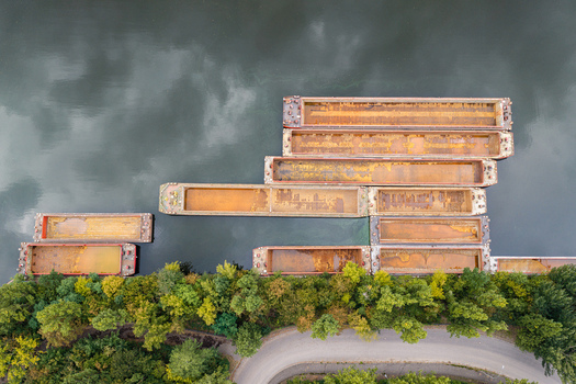Aerial view of several rusted cargo barges moored along a riverbank lined with lush green trees and a winding road in Berlin Berlin Germany.