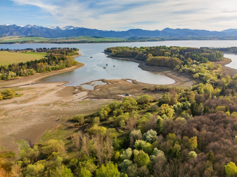 Aerial view of Liptovska Mara reservoir with lush green forests sandy shores and the snow capped Tatra Mountains under a blue sky Liptovsky Trnovec Zilina Region Slovakia.