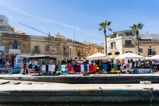 View of a vibrant outdoor market with colorful t shirts on display along the waterfront promenade under a clear blue sky in Marsaskala Southern Region Malta. View of a vibrant outdoor market with colorful t shirts on display along the waterfront promenade under a clear blue sky in Marsaskala Southern Region Malta.