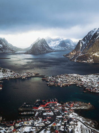 Aerial view of Reinebringen s village nestled amid frigid waters jagged snow capped peaks under brooding skies Reinebringen Nordland Norway.