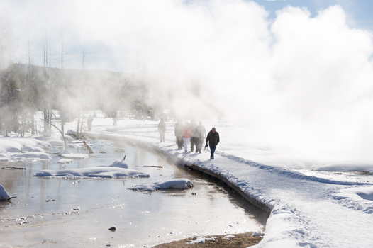 View of people walking along a snow covered boardwalk through a geothermal area with rising steam in Yellowstone National Park Wyoming United States. View of people walking along a snow covered boardwalk through a geothermal area with rising steam in Yellowstone National Park Wyoming United States.